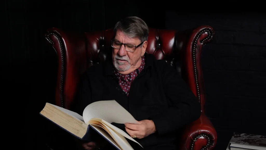 Carl Chinn MBE seated in a red leather chair